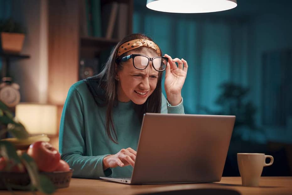 A nearsighted woman squints at her laptop screen while holding her reading glasses away from her face